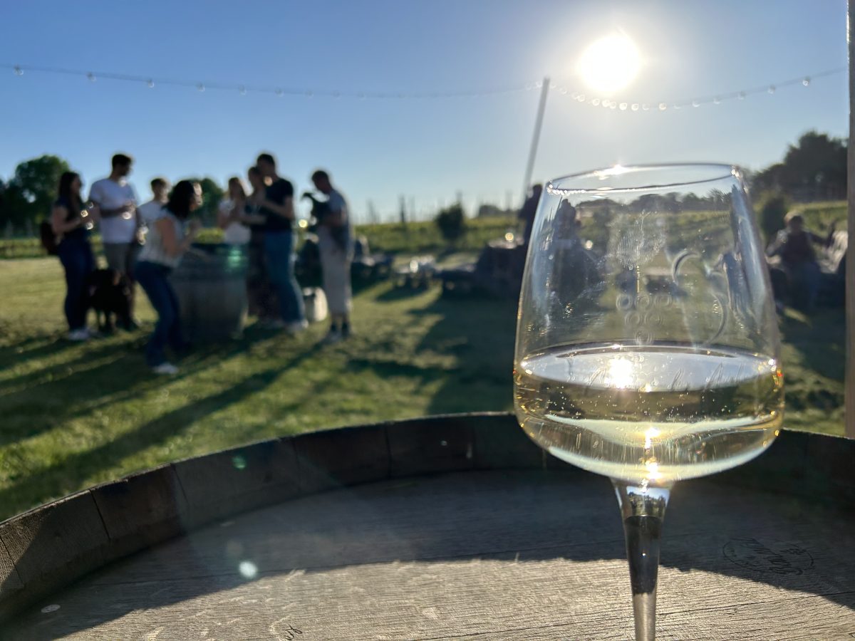 Un verre de vin blanc posé sur un tonneau, un groupe en contre-jour à l’arrière-plan